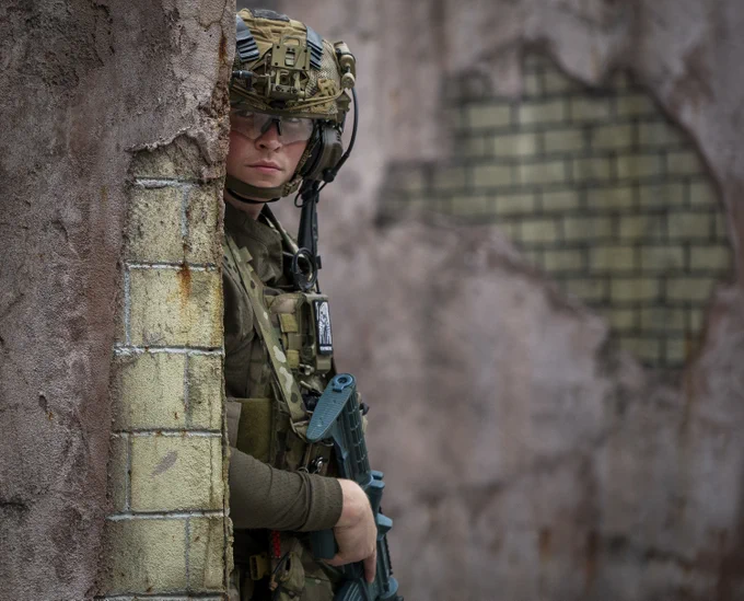 Airman in tactical gear and helmet holding a rifle, partially hidden behind a weathered brick wall in an urban setting.