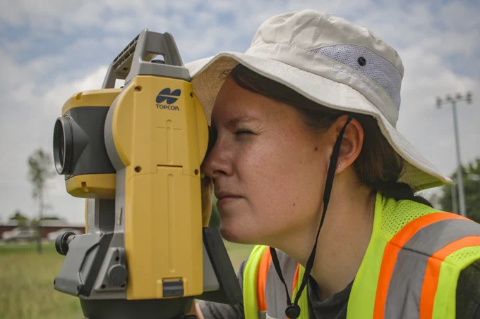 A student in a construction vest and sun hat peeps through a yellow topographical surveyor, a Total Station.
