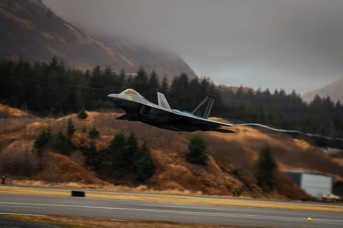 Military jet flying low over a runway with forested hills in the background under a cloudy sky.