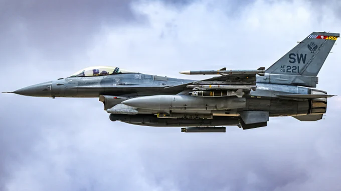 F-16 fighter jet flying against a cloudy sky with visible missiles and external fuel tanks attached.