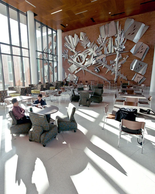 An open atrium space with chairs and study areas. In the background on a brick wall is an abstract metal sculpture that also continues outdoors through the glass windows.