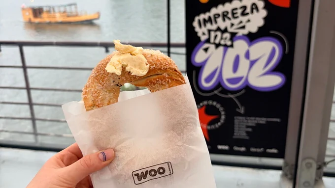 A hand holds up a pastry in a paper bag with the Woo logo on it in front of the views of a river with a boat and a sign for the Woo Community Event at WCEU 2026.