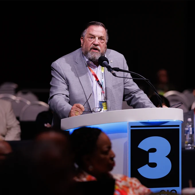 IATSE President Matt Loeb in a gray blazer, speaking at a podium marked with a large number 3 during a conference or event.