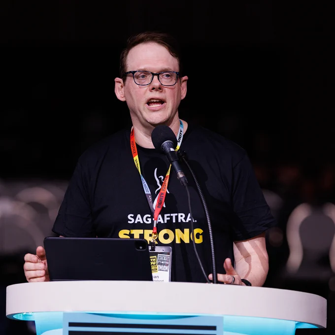 SAG-AFTRA National Executive Director & Chief Negotiator Duncan Crabtree-Ireland, wearing a black 'SAG AFTRA STRONG' shirt, speaking at a podium with a microphone and tablet in front of an empty auditorium.