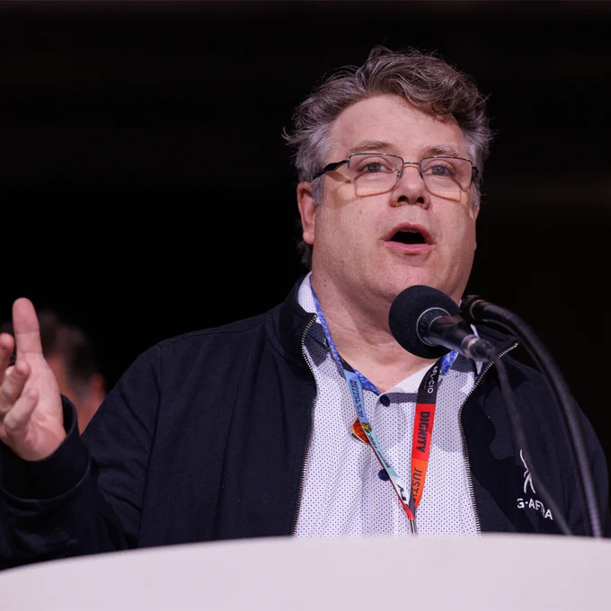 SAG-AFTRA President Sean Astin is wearing a black jacket and white shirt, speaking into a microphone at a podium during an event.