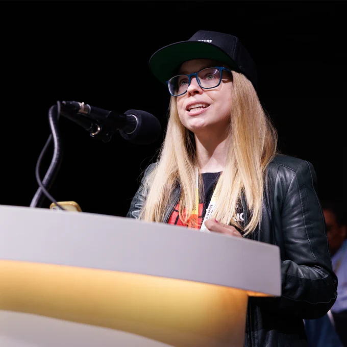 Writers Guild of America East Secretary-Treasurer Sasha Stewart, with long blonde hair wearing a black cap and leather jacket, speaking at a podium with a microphone on stage.