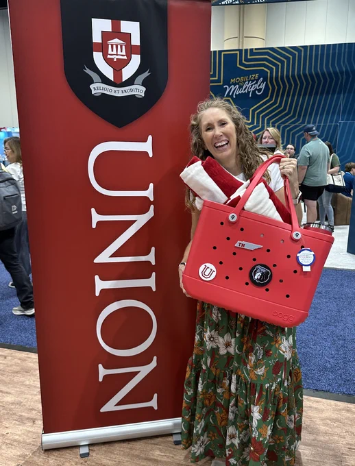 Smiling woman holding a red tote with a folded red-and-white towels beside a red university banner at a busy booth.