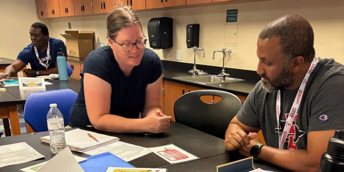 Three adults engaged in a classroom setting with papers and water bottles on the table, focused on discussion or study.