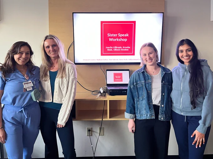 A group of four people standing in front of a wall-mounted screen displaying a slide titled "Sister Speak Workshop" with names listed below. Two individuals wear casual clothing, one wears scrubs, and one is in business attire. A wooden shelf holds a laptop beneath the screen.