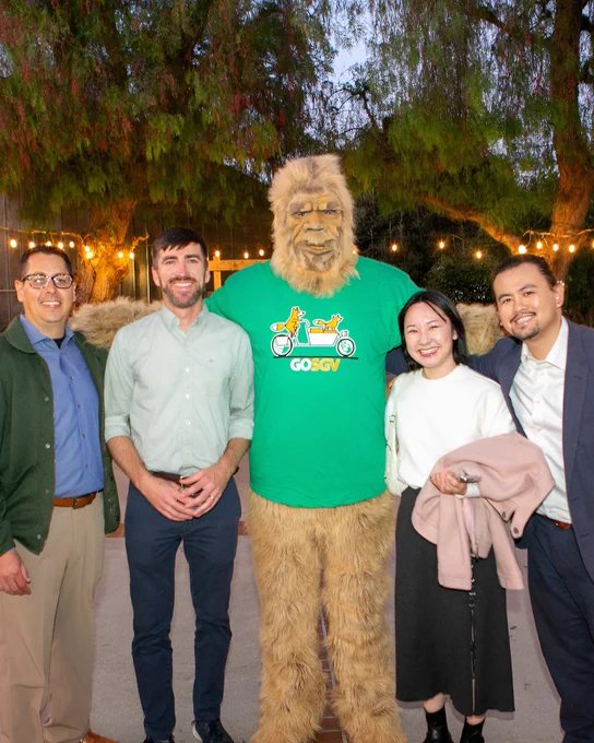 A group of five people stands outdoors on a paved area under string lights. In the center is a person in a Bigfoot costume wearing a green "GoSGV" T-shirt. The others are dressed in semi-formal attire, smiling for the camera, with trees in the background.