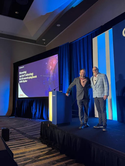 Two Siteimprove speakers stand on a Gartner conference stage beneath a large screen showing the session title "Showing up isn't winning: An AI search playbook with Ryder," with the Siteimprove logo visible in the lower left corner.