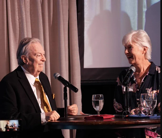 Smiling man and woman sit at a table with microphones. A mobile device is seen in the foreground recording the conversation.