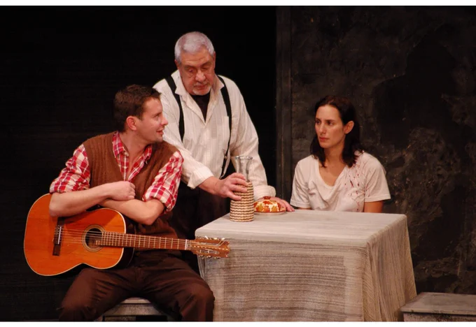 Fotografía de una escena teatral con tres personas:  un hombre con guitarra sentado, otro de pie sirviendo bebida y una mujer sentada frente a una mesa con mantel.