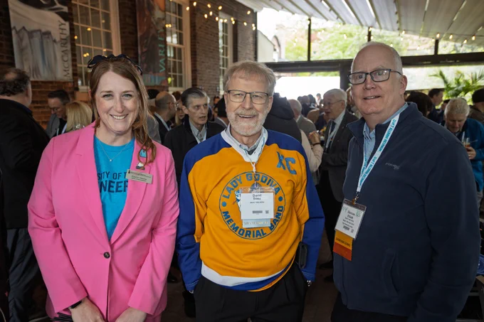 Three people stand together at a crowded indoor reception, smiling toward the camera. One is wearing a Lady Godiva Memorial Band jersey. Framed artwork banners and string lights hang behind them.