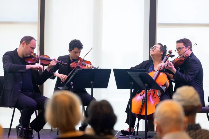 The Calidore String Quartet performs on stage, featuring two violinists, a violist, and a cellist. They are focused on their music, and several people in the audience watch attentively in the foreground.