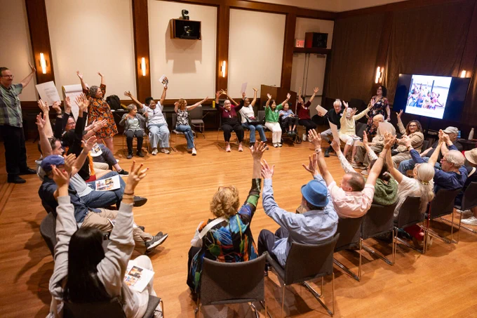 A group of people seated in a circle in a room, all raising their hands enthusiastically. A screen displays a colorful image, and various papers are held by participants.