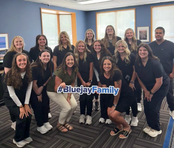 Group of students in dark medical scrubs posing indoors in a classroom setting. A few individuals hold a “#BluejayFamily” sign, with windows and framed wall art in the background.