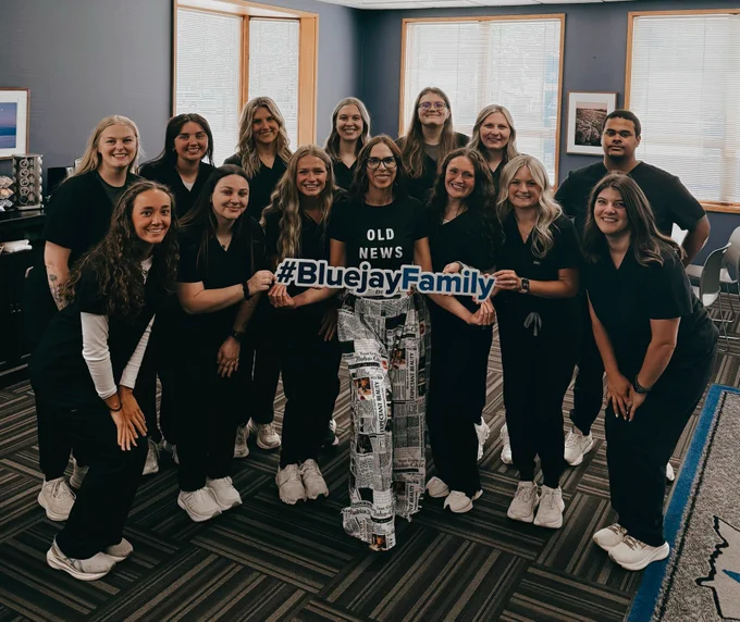 Group of students in dark medical scrubs gathered indoors in a classroom with blue walls and large windows. Several hold a cutout sign reading “#BluejayFamily.”