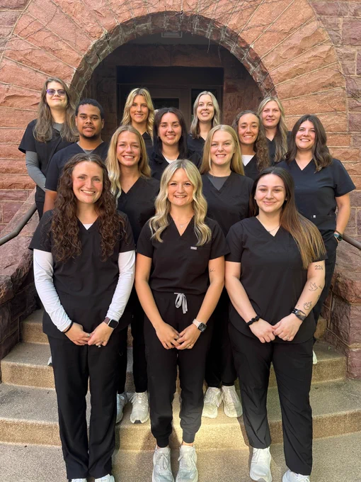 Group of students in dark medical scrubs standing on outdoor steps in front of a stone building with a large arched doorway. The group is arranged in rows, wearing black uniforms and white shoes, with natural light highlighting the stone texture and entryway behind them.