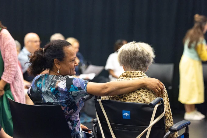 A person affectionately places an arm around another seated in a wheelchair, creating a warm and caring atmosphere. Several others are present in the background, engaging in conversation.
