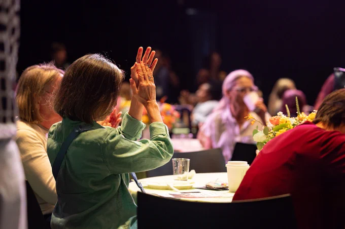 A group of people seated at tables, some clapping and others interacting, with floral centerpieces visible. The lighting is dim, creating a focus on the individuals.