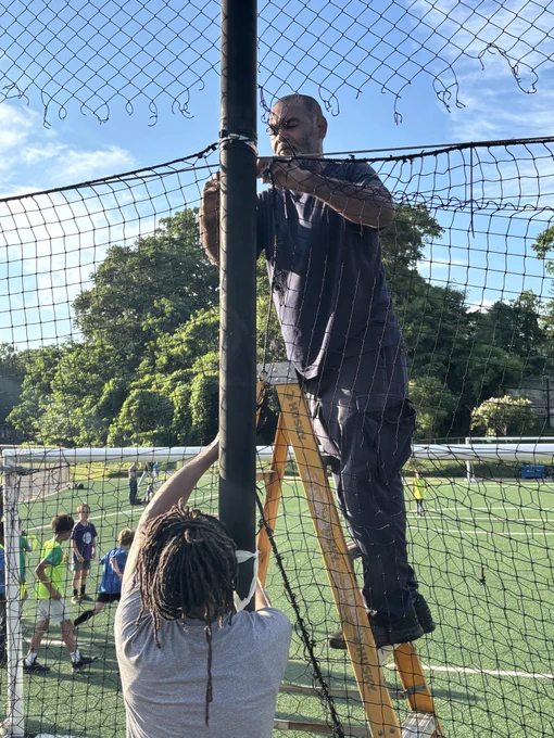 Two men fixing a torn net on a sports field, one standing on a ladder and the other holding the pole steady during a sunny day.