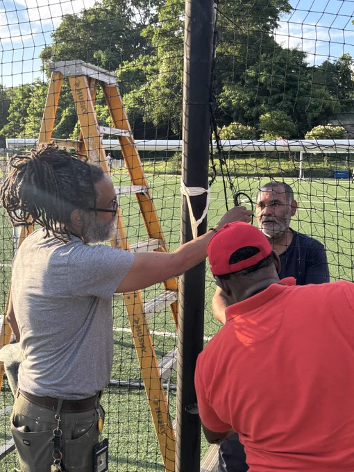 Three people working together to install or repair a net on a sports field with a ladder nearby under a sunny sky.