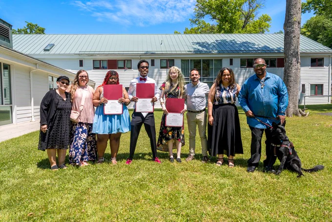 Group photo of eight people standing outside the VRC campus, 3 of whom hold up certificates of completion. 