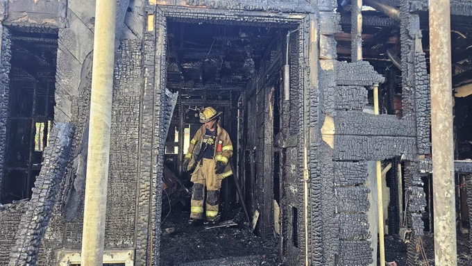 A firefighter stands inside a heavily charred and partially destroyed structure, assessing the aftermath of a fire. The walls and ceiling are blackened with soot and ash. The firefighter is wearing protective gear and a helmet, carrying equipment.