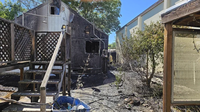 A charred and blackened wooden house stands with visible fire damage. The exterior features several broken windows and partially burnt walls, with nearby trees and vegetation showing signs of burning. A wooden staircase leads up to a deck on the side of the house. The ground is littered with debris. Clear blue sky and trees are visible in the background.