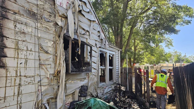 A fire-damaged house with charred and melted siding is surrounded by yellow caution tape. Burnt debris is visible near the structure. An individual in safety gear, wearing a construction vest, walks near the scene. Trees and a partially visible fire truck are in the background.