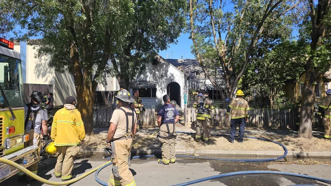 Emergency personnel in firefighting gear stand in front of a residential house with visible smoke damage. A firetruck is parked on the street, and hoses are deployed. Trees and a fence are in the foreground.