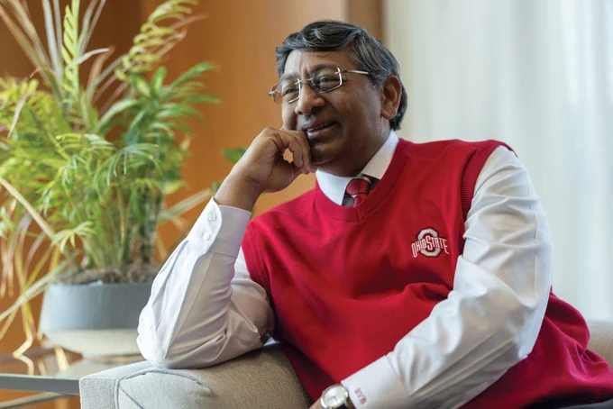 Ravi Bellamkonda leans one elbow on the arm of a couch as he leans into it and chuckles. He wears a scarlet Ohio State sweater vest, glasses and has salt-and-pepper tussled hair.