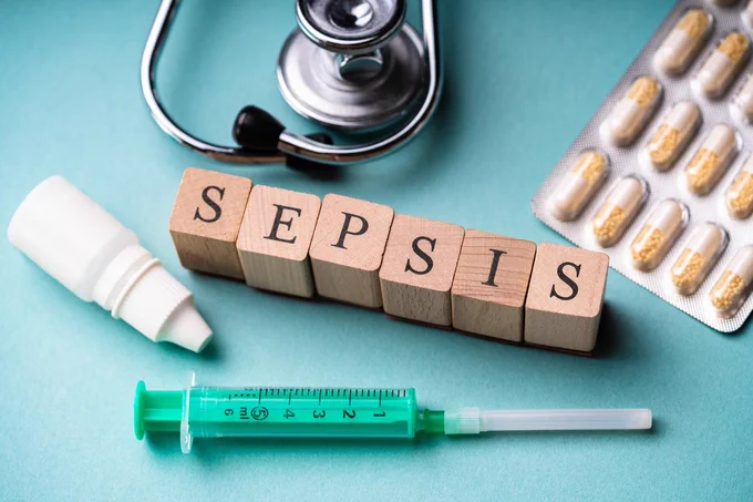 Sepsis concept photo: Wooden blocks spelling 'SEPSIS' surrounded by a stethoscope, spray, syringe, and blister pack of capsules on a blue surface.