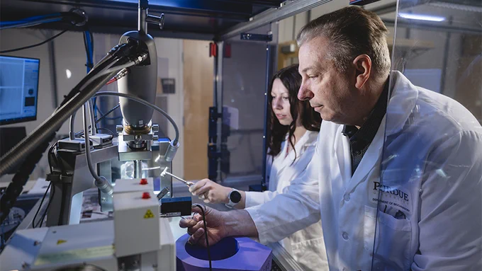 Two people in a laboratory setting are working with advanced equipment. They are wearing white lab coats with "Purdue" logos. Various screens, wires, and instruments are visible around them, indicating a scientific research environment.