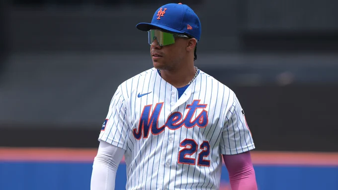 Baseball player Juan Soto of the New York Mets. He walks on the field with a determined expression on his face, his eyes hidden by reflective sunglasses. He wears a white pinstripe uniform, with “Mets” and “22” emblazoned across the front, along with a blue hat. The stadium is out of focus behind him. 
