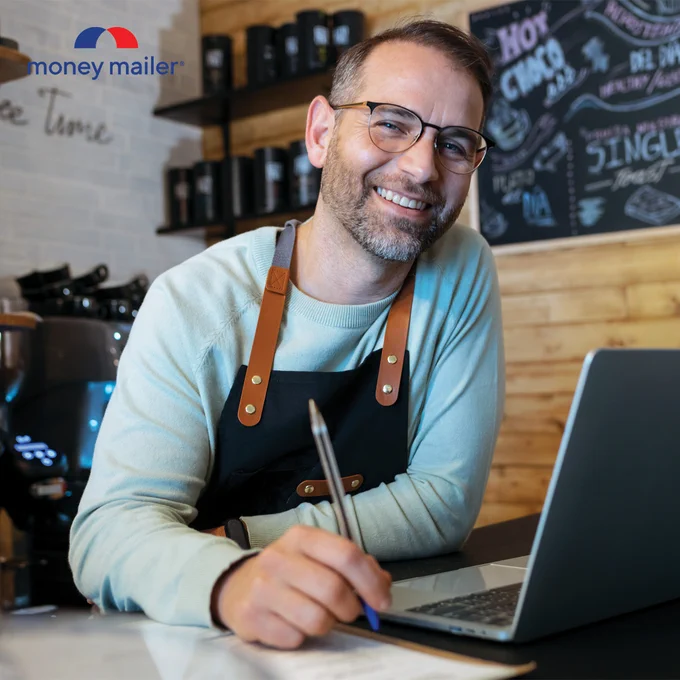 Barista in a light blue sweater and black apron writing with a pen while working on a laptop in a cozy coffee shop.