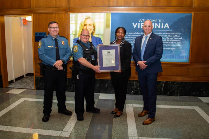 Four officials stand indoors in front of a Virginia welcome sign, with one woman holding a framed certificate.