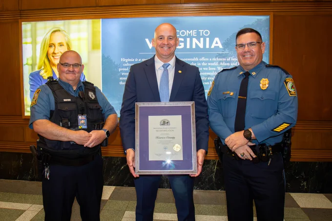 Three officials stand indoors in front of a 'Welcome to Virginia' sign, with the central figure holding a framed certificate.