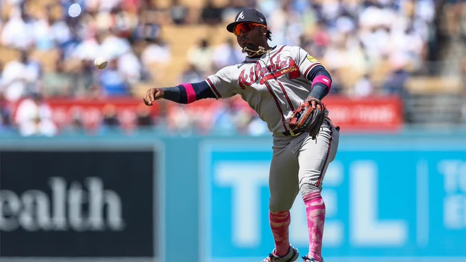 Baseball player Ozzie Albies of the Atlanta Braves. He is in motion on the field, throwing a baseball with a focused expression on his face. He wears a gray Braves jersey with "Atlanta" displayed across the front, matching pants, red reflective sunglasses, and a navy blue hat. Behind him, spectators in the stadium are out of focus.