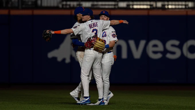 Baseball player Carson Benge of the New York Mets. He stands on the field with his back to the camera, embracing two of his teammates in a celebratory hug. He wears a white pinstripe Mets jersey with his last name and the number 3 emblazoned across the back, and matching pants. Behind them, a dark blue stadium wall is visible in the background.
