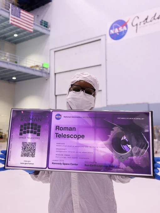 A person in a head-to-toe white clean room suit holds a large purple "boarding pass" for NASA's Nancy Grace Roman Space Telescope in Goddard's large cleanroom. The boarding pass includes an image of the 18 detectors, an artist concept image of the telescope, a QR code, and the NASA logo. "Roman Telescope" is written where the name would go.