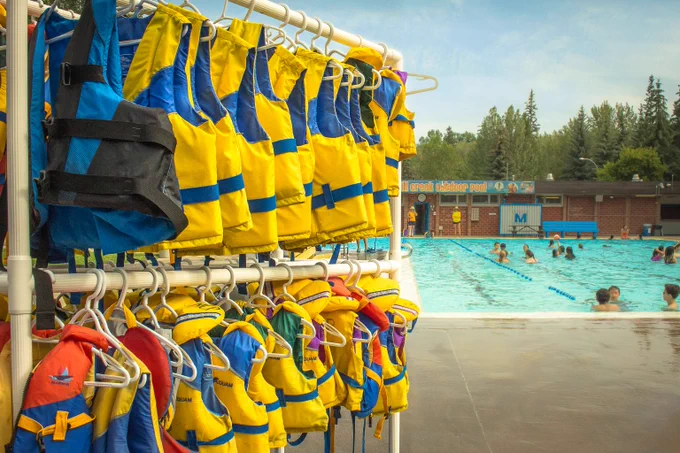 Bright yellow and blue life jackets hanging on racks beside an outdoor swimming pool with swimmers and trees in the background.