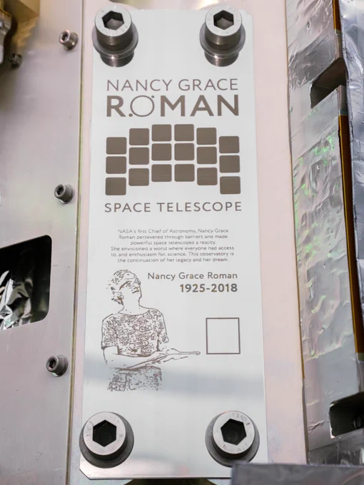 The plate attached to the Roman telescope, where a memory card containing names will be attached. It's a tall, thin, rectangular silver plate. At the top of the plate is the stylized Nancy Grace Roman name logo. Below is the silhouette of Roman's 18-square detectors in their iconic arch shape. Text below that reads "NASA's first chief astronomer, Nancy Grace Roman, persevered through barriers and made powerful space telescopes a reality. She envisioned a world where everyone had access to, and enthusiasm for, science. This observatory is the continuation of her legacy and dream." Below the text is an illustration of Dr. Roman looking up, next to her name and the years 1925-2018. Below the years is a small box where the memory card will be affixed. The plate is held down with two large bolts at the top and bottom.