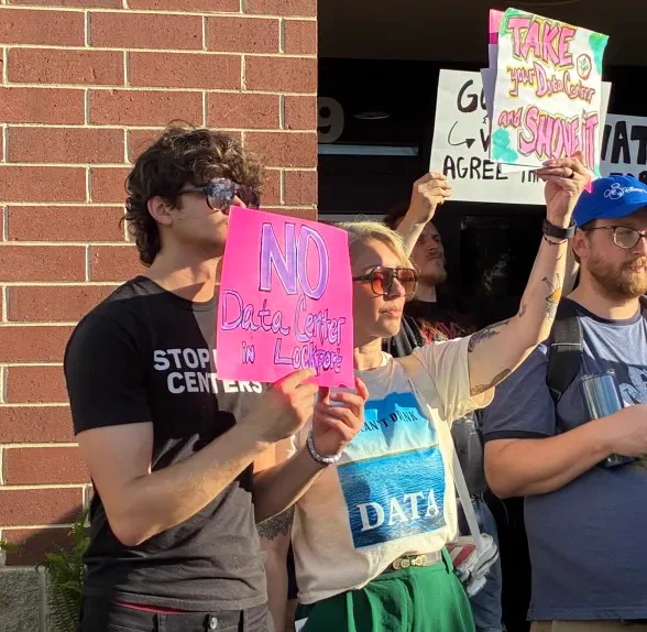 Protesters holding signs against data centers, one reading 'NO Data Centers in Lockhart' in front of a brick wall.