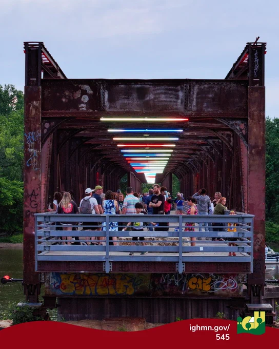 Group of people gathered on the Rock Island Swing Bridge adorned with colorful LED lights overhead during dusk.
