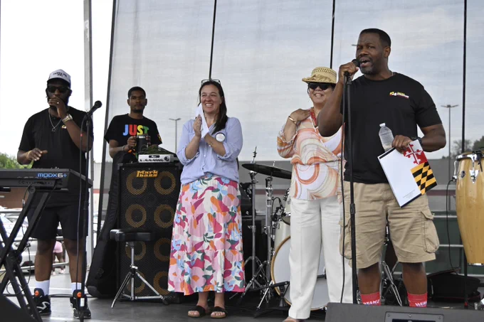 Group of five musicians performing on an outdoor stage with various instruments and microphones.