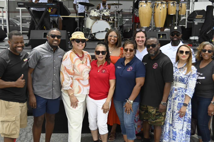 Group of diverse adults posing together in front of a stage with musical instruments during an outdoor event.