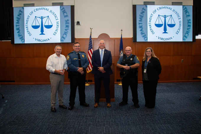 Five officials stand in a conference room with two screens displaying the Virginia Department of Criminal Justice Services logo behind them.
