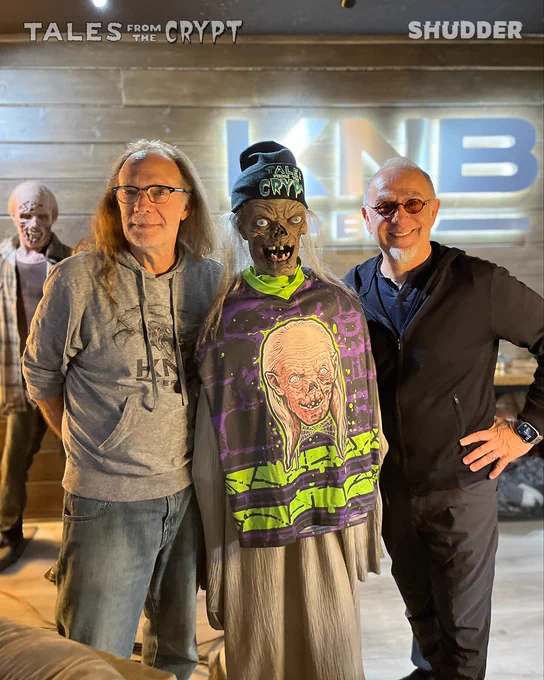 Two men stand beside a Crypt Keeper figure in a setting featuring 'Tales from the Crypt' branding. The mood is nostalgic and amusing.